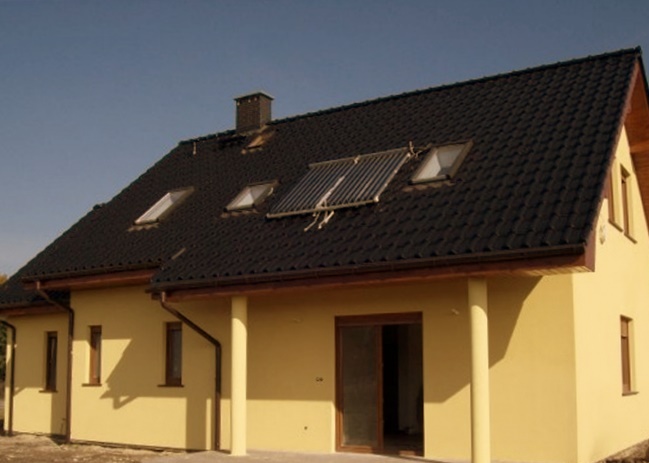 Żółta elewacja domu jednorodzinnego z ciemnym dachem, na którym zamontowane są kolektory słoneczne i okna dachowe.